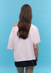 Světle růžové oversized tričko s designem spadlých ramenních švů, z bavlněného materiálu a krátkými rukávy, doplněné černou sukní.