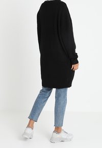 Czarny, oversize sweter z dzianiny, o miękkiej fakturze, z długimi rękawami, luźny krój, noszony na jasnoniebieskich przetartych dżinsach i szarych butach sportowych.