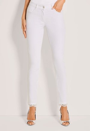 Frau trägt schmale weiße Jeans mit Spitzenbesatz an den Knöcheln und silberne offene Absatzschuhe, steht vor einem einfarbigen hellen Hintergrund.