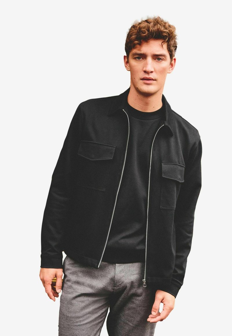 Black jacket with a collar, front zipper, two chest pockets, made of smooth fabric. Worn over a black shirt and gray trousers.