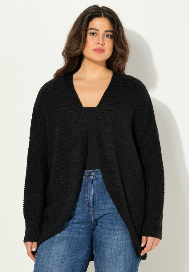 Svart stickad cardigan med öppen front, vida ärmar och avslappnad passform. Bärs över en svart topp och kombineras med blå jeans.