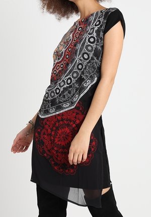 Kobieta ubrana w czarną sukienkę z czerwono-szarymi, misternie zdobionymi wzorami mandali, do tego czarne, sięgające uda kozaki oraz bransoletka na nadgarstku.