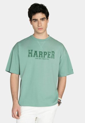 Hombre joven con una camiseta verde holgada con el texto "HARPER" y pantalones blancos, de pie frente a un fondo claro y liso.