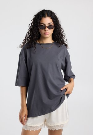 Vrouw met krullend haar die donkere zonnebril draagt, een loszittend donkergrijs t-shirt, witte shorts met kantafwerking en een gouden ketting tegen een effen achtergrond.
