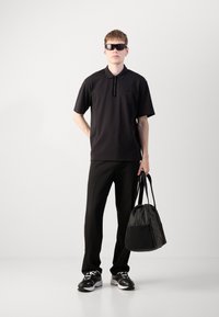 Svart polo-skjorte med glidelåskrage, svarte bukser og svarte sneakers. Holder en svart stoffbag med rund bunn og korte stropper.