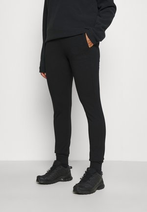 Person trägt schwarze, eng anliegende Jogginghosen, schwarzen Pullover und schwarze Sportschuhe und steht vor einem schlichten hellen Hintergrund.