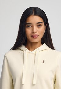 Sudadera crema con un bolsillo canguro frontal, capucha con cordón y una pequeña figura bordada en el pecho. Textura de tela suave.