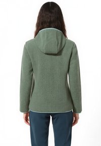 Donna con capelli lunghi che indossa una giacca di pile verde con cappuccio e pantaloni blu scuri, vista di spalle su uno sfondo bianco.