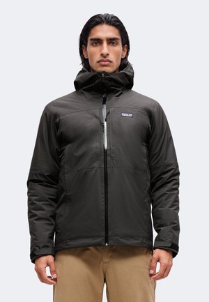Mand med mørkt hår iført en sort Patagonia hættejakke, lynet helt op, kombineret med beige bukser, stående mod en ensfarvet lys baggrund.