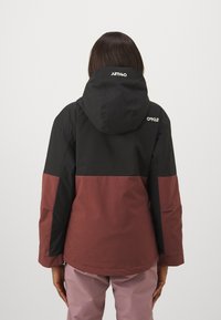 Veste imperméable avec une section supérieure noire et une section inférieure bordeaux. Comprend une capuche et des poches zippées. Texture lisse.