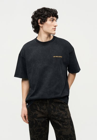 Νέος άνδρας με σγουρά μαλλιά που φοράει μαύρο t-shirt "LES BENJAMINS" και σκούρο παντελόνι με λουλουδάτο μοτίβο, κοιτάζοντας προς τα πλάγια.