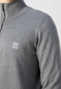 Gros plan d'une personne portant un pull gris à fermeture éclair avec un col côtelé et un petit logo carré BOSS sur la poitrine.