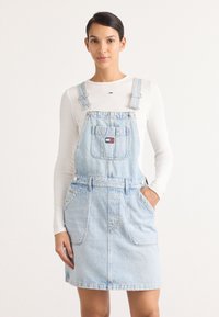 Lys blå denim overallkjole med justerbare stropper, frontlomme og sidespenner. Båret over en langermet hvit skjorte.