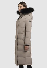 Lang beige pufferjakke med en hætte beklædt med pels, quiltet design, sidelommer og et mærket patch på venstre arm. Skræddersyet pasform.