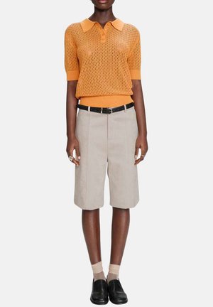 Femme portant un polo en maille orange à manches courtes, un short beige à hauteur du genou avec une ceinture noire, des chaussettes transparentes beige et des chaussures noires sans lacets.
