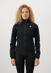 Žena s curls blondinom koja nosi crnu sportsku duksericu s patentnim zatvaračem i crne biciklističke hlače, stoji pred jednostavnom bijelom pozadinom.