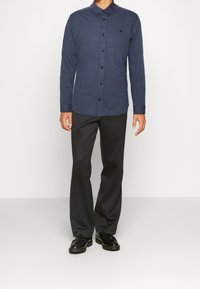 Knappupp skjorta i mörkblått tyg med långa ärmar, som har en krage, bröstlogotyp och matchande knappar, i kombination med mörka byxor och svarta skor.