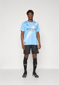 Svetlo modra nogometna majica z belimi diagonalnimi poudarki, črne kratke hlače, črne nogavice in nogometni copati. Na majici je logotip ETIHAD AIRWAYS.