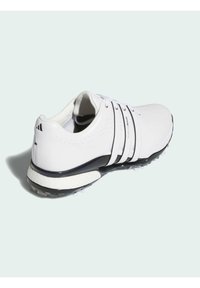 Bílé golfové boty s třemi černými pruhy, perforovaným horním dílem pro ventilaci a texturovanou podrážkou s viditelným vzorem hrotů.