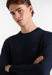Suéter azul marino con cuello redondo, puños acanalados y un pequeño logo azul en el pecho. Textura suave y diseño ajustado.