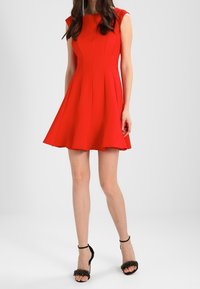 Czerwona sukienka w kształcie litery A z dopasowanym górą i rękawami typu cap, z plisami, zestawiona z czarnymi sandałami na szpilkach ozdobionymi dekoracyjnymi akcentami.
