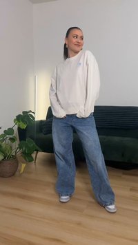 White sweatshirt with light blue logo, oversized fit; blue wide-leg jeans; gray sneakers; indoor setting with green plant and sofa.