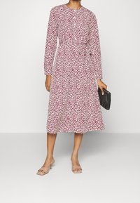 Donna che indossa un abito midi bianco con stampa astratta rossa, vita legata, maniche lunghe, sandali con tacco color beige e in mano una pochette nera.
