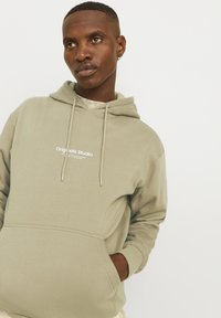 Olijfgroene hoodie van zachte stof, met een voorkeurszak, een capuchon met trekkoord en een wit "Originals Studio" logo op de borst.