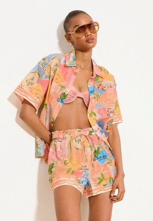 Kobieta nosząca oversize wielokolorową koszulę zapinaną na guziki oraz pasujące szorty na różowym topie bikini, z dużymi brązowymi okularami przeciwsłonecznymi.