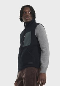 Gilet en polaire de couleur bleu marine foncé avec un pull gris, comportant une poche poitrine verte, une fermeture éclair et des poches latérales. Tissu texturé avec des accents lisses.