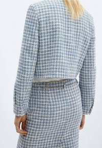 Donna con capelli biondi che indossa un giubbotto corto in tweed a scacchi blu e bianchi e una gonna coordinata, vista da dietro.