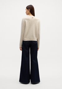Beige gebreide trui met een gestructureerde afwerking, voorzien van een ronde halslijn en ribgebreide manchetten, in combinatie met donkerblauwe wijde jeans.