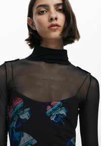 Turtleneck in mesh nero con motivi floreali in blu e rosa, indossato sopra un vestito attillato con scollatura profonda e spalline delicate.