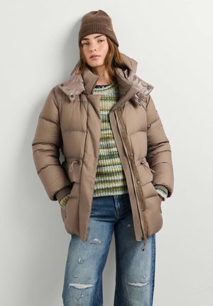Jonge vrouw met een bruine beanie, beige donsjas, gestreepte trui en gescheurde blauwe spijkerbroek staand tegen een eenvoudige lichte muur.