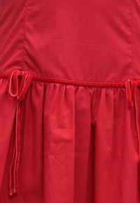 Červené šaty s nabíranou sukní, s hladkou texturou a sametovými pásky v oblasti pasu. Jednoduchý design v jednotné barvě.