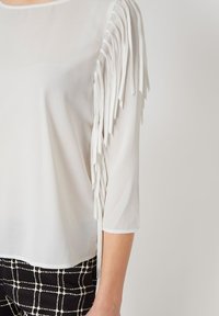 Blusa blanca con mangas tres cuartos que presenta detalles de flecos en el hombro izquierdo. La tela es transparente con una textura suave.