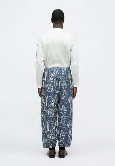 Λευκό μπλουζάκι με μακριά μανίκια, μπλε παντελόνι φαρδιάς γραμμής με σχέδια που μοιάζουν με άσπρα ζωγραφισμένα σκίτσα, και καφέ παπούτσια.