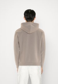 Hombre de espaldas vistiendo una sudadera color topo y pantalones blancos frente a un fondo blanco liso.