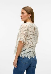 Nėrinių blusa off-white spalvos, pabrėžianti gėlių raštus, su banguotais rankovėmis ir detale raktui nugarinėje pusėje. Derinama su mėlynomis džinsomis.