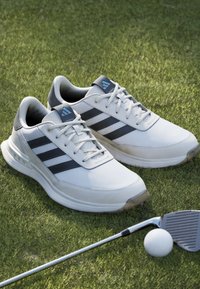 Vita golfskor med grå detaljer och tre mörka ränder. Tillverkade av syntetiskt material, med en vadderad krage och en strukturerad sula.