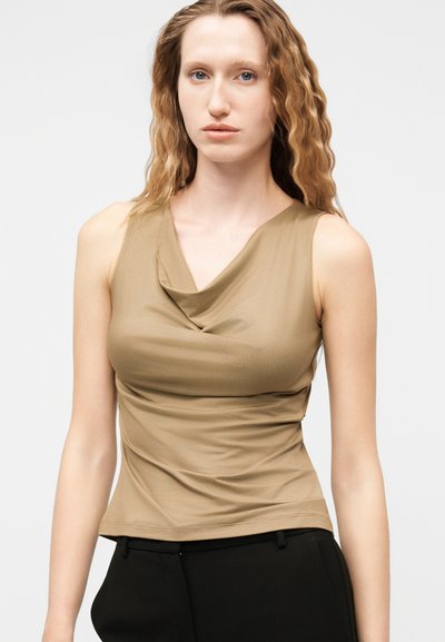 Γυναίκα με μακριά κυματιστά μαλλιά, ντυμένη με αμάνικο μπεζ τοπ με ντραπέ λαιμόκοψη και μαύρο παντελόνι, που κοιτάζει προς τα εμπρός σε απλό φόντο.