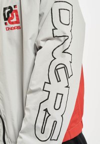 Light gray jacket with red accents, featuring large black lettering on the sleeve and a logo on the chest. Smooth texture and casual design.