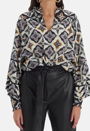 Kvinde iført sorte læderbukser med høj talje og bindebælte samt en mønstret bluse med lange ærmer og geometriske designs.