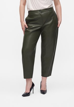 Vrouw draagt korte donkergroene leren broek, witte top en zwarte hoge hakken, staat met handen in haar zakken.