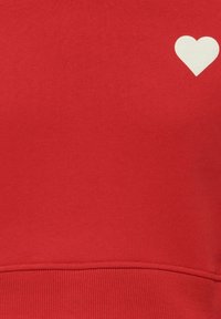 Punainen huppari, joka on valmistettu pehmeästä kankaasta ja jossa on pieni valkoinen sydänkuvio vasemmalla ylhäällä. Suuri, sileä rakenne, jossa on ribbireunus.