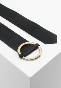Svart lærbelte med en sirkulær gullspenne. Har en jevn tekstur og en avsmalnende ende for et elegant design.