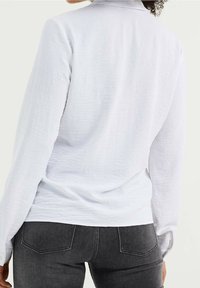 Bílá košile s dlouhým rukávem z texturované látky, s vysokým límcem a bočními rozparky, v kombinaci s tmavě šedými úzkými džíny se zadními kapsami.