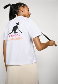 Valge puuvillane t-särk, millel on must kangaruugraafika, kes hoiab golfikeppi, ja roosa ning oranž tekst, millel on kirjas "KANGOL" ja "J. LINDEBERG."