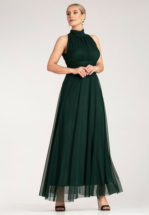 Langes, dunkelgrünes Kleid mit hohem Ausschnitt, tailliertem Oberteil, schwingendem Rock, geschichtetem Organza und sichtbaren modernen Absätzen.