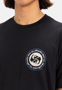 Camiseta de algodón negra con un logo circular en negro, blanco y amarillo, situado en el área izquierda del pecho. Diseño clásico de cuello redondo.